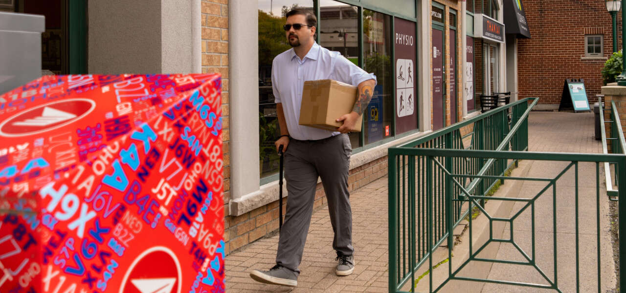 A man carrying a package walks with an assistive cane on a sidewalk ramp along storefronts. 