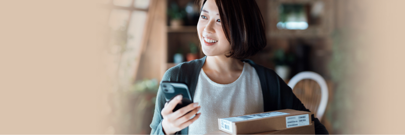 A smiling woman holds a smartphone in 1 hand and shipping packages under her other arm.