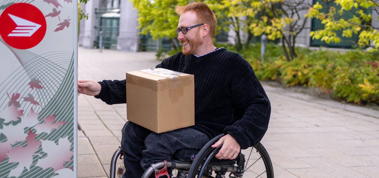 Un homme en fauteuil roulant ramasse un colis dans une boîte postale communautaire de Postes Canada.