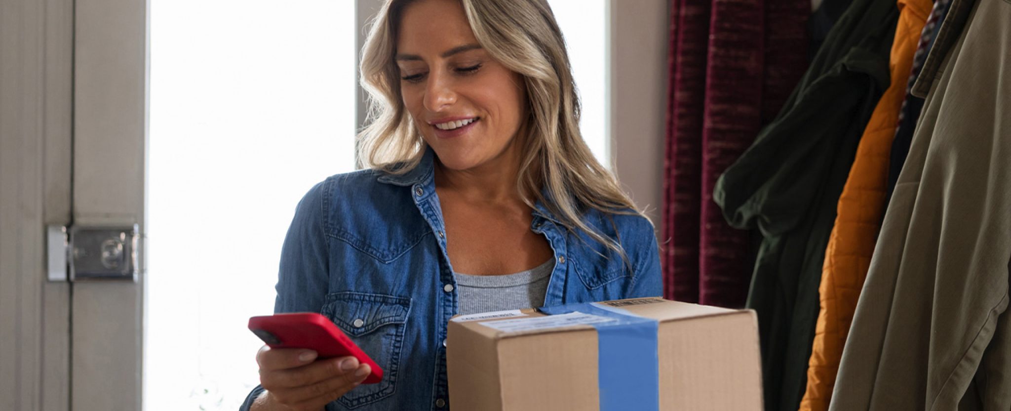 Une femme souriante regarde son téléphone intelligent en tenant un colis.