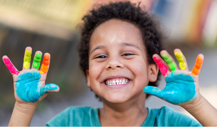 Un jeune enfant sourit en levant les mains, chaque doigt et chaque paume étant peints d'une couleur différente.