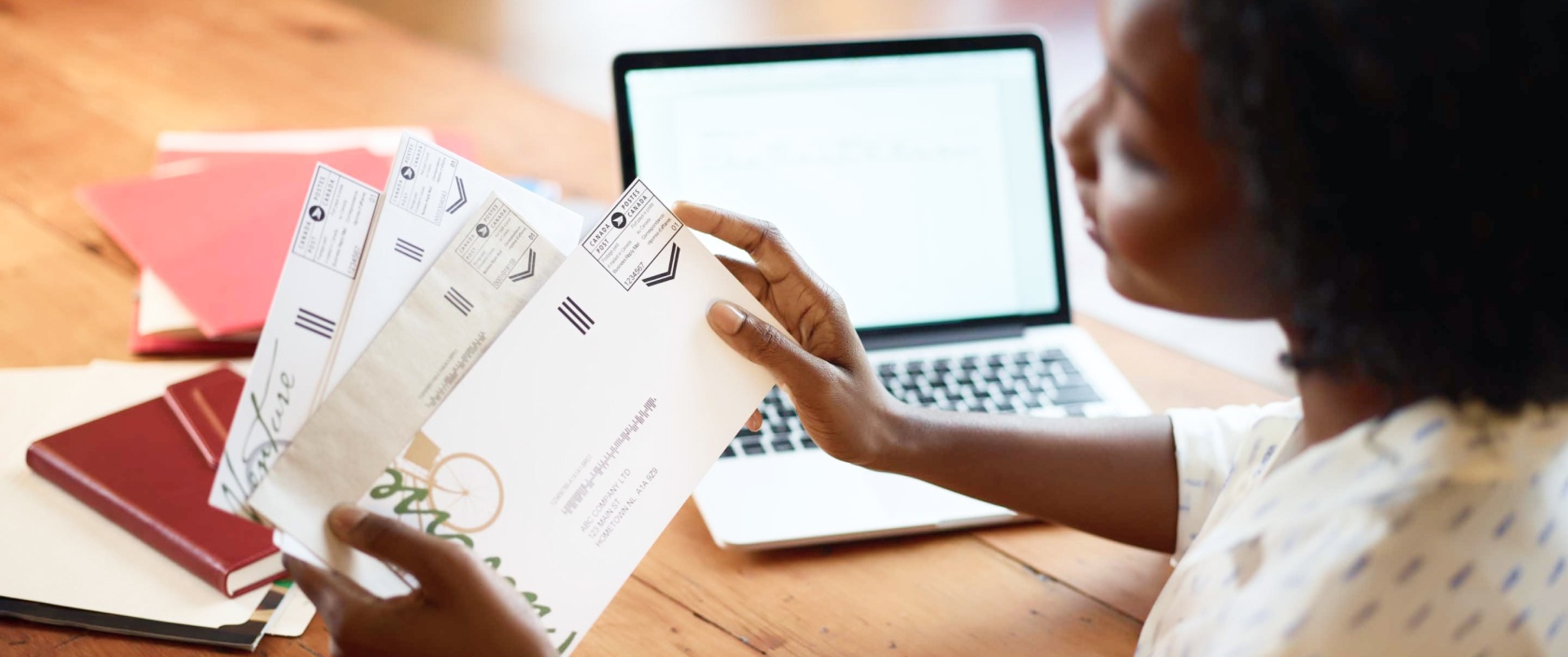 Une femme à son bureau tient du courrier d’affaires. Il y a une vignette postale de Postes Canada dans le coin supérieur droit de chaque enveloppe.