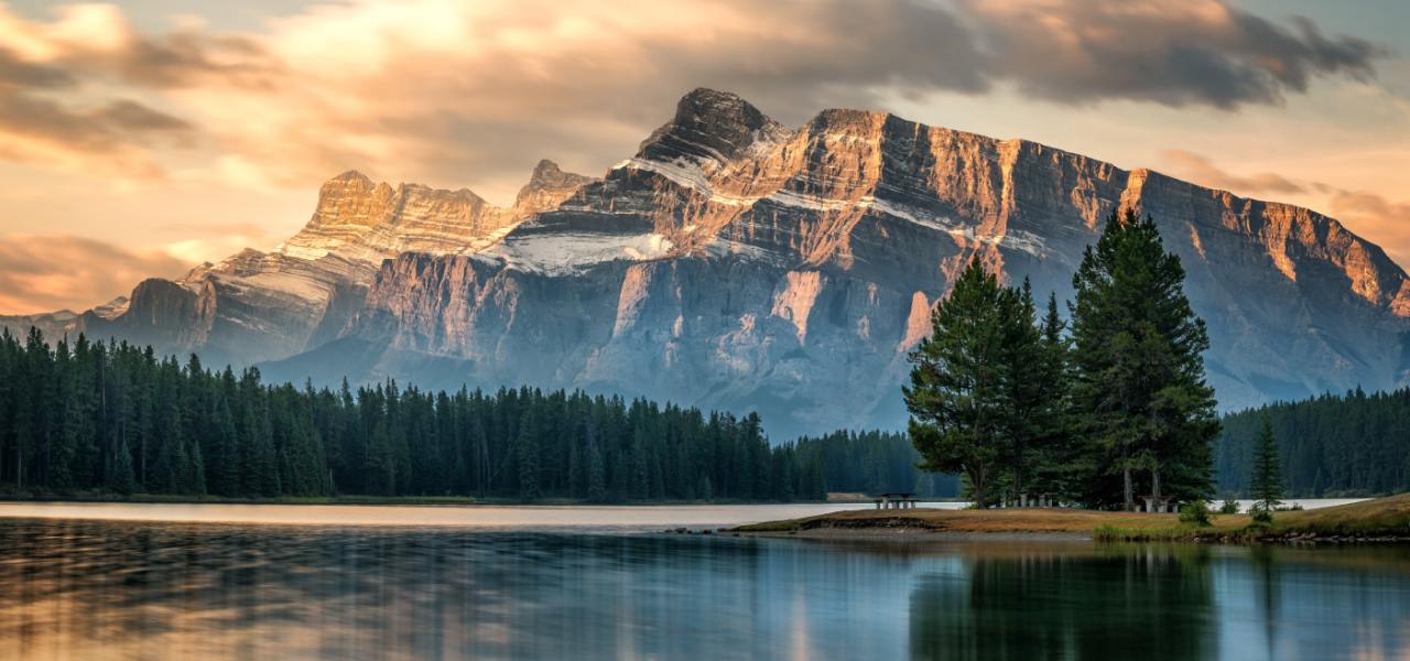 Un lac paisible entouré d’une forêt qui se situe au pied d’une montagne rocheuse et qui est baigné dans la lumière dorée d’un coucher de soleil.