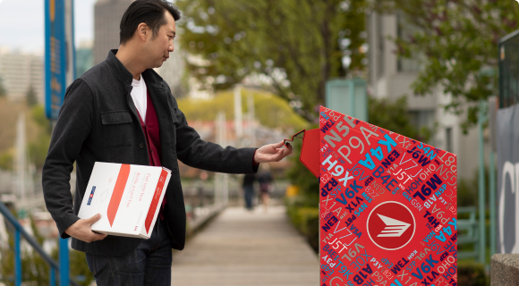 Un homme sur un trottoir ouvre la chute d’une boîte aux lettres publique de Postes Canada. Il tient une boîte à tarif fixe dans son autre main.